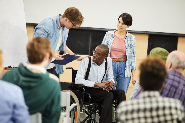 Young man in glasses supporting another man in a wheelchair while at the front of a conference.